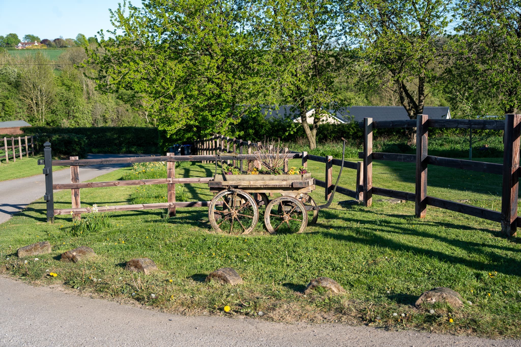 An old wooden farm cart planted up on the lawn at Newbound Farm, framed by dark post-and-rail fencing, blossoming trees and open Nottinghamshire countryside beyond.