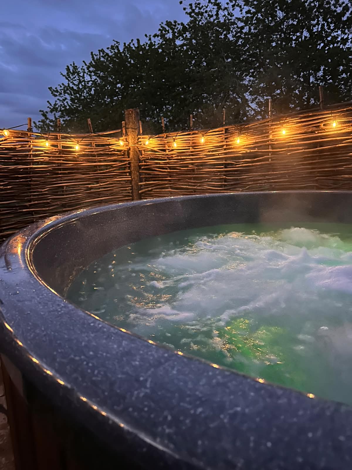 The hot tub bubbling after dark, steam drifting off the lit water, with warm festoon bulbs strung along the willow screen against a deep blue evening sky.