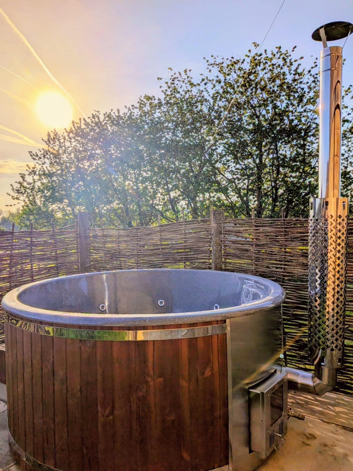 The hot tub at sunset, the low sun flaring through bare trees behind the willow screen and catching the stainless steel flue of the wood burner that heats it.