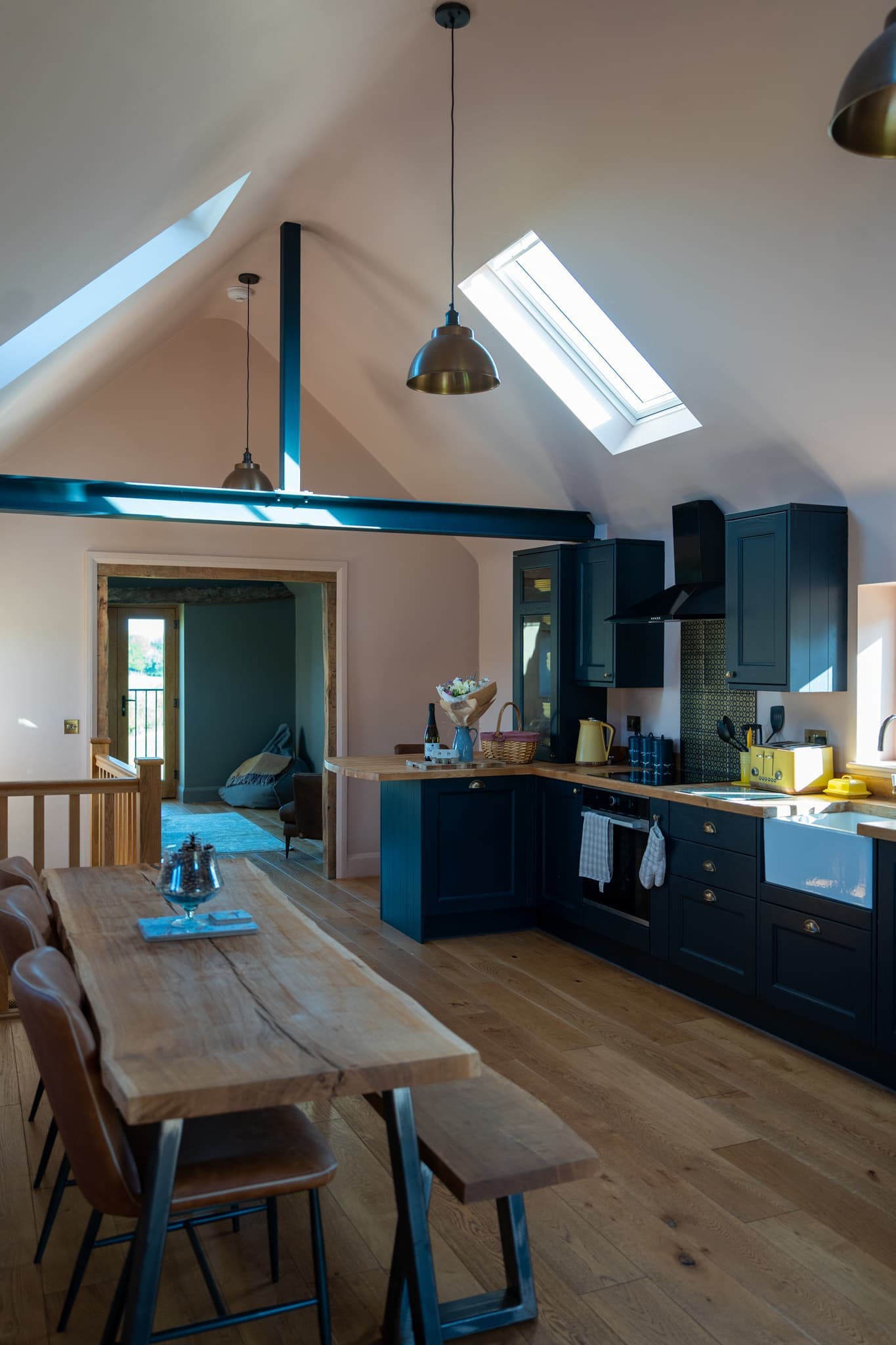 The open-plan kitchen and dining area under a vaulted ceiling with two rooflights: deep navy shaker cabinets, a butler sink, a long live-edge oak table with leather chairs and a bench, and brass pendant lights hanging from a painted steel beam.