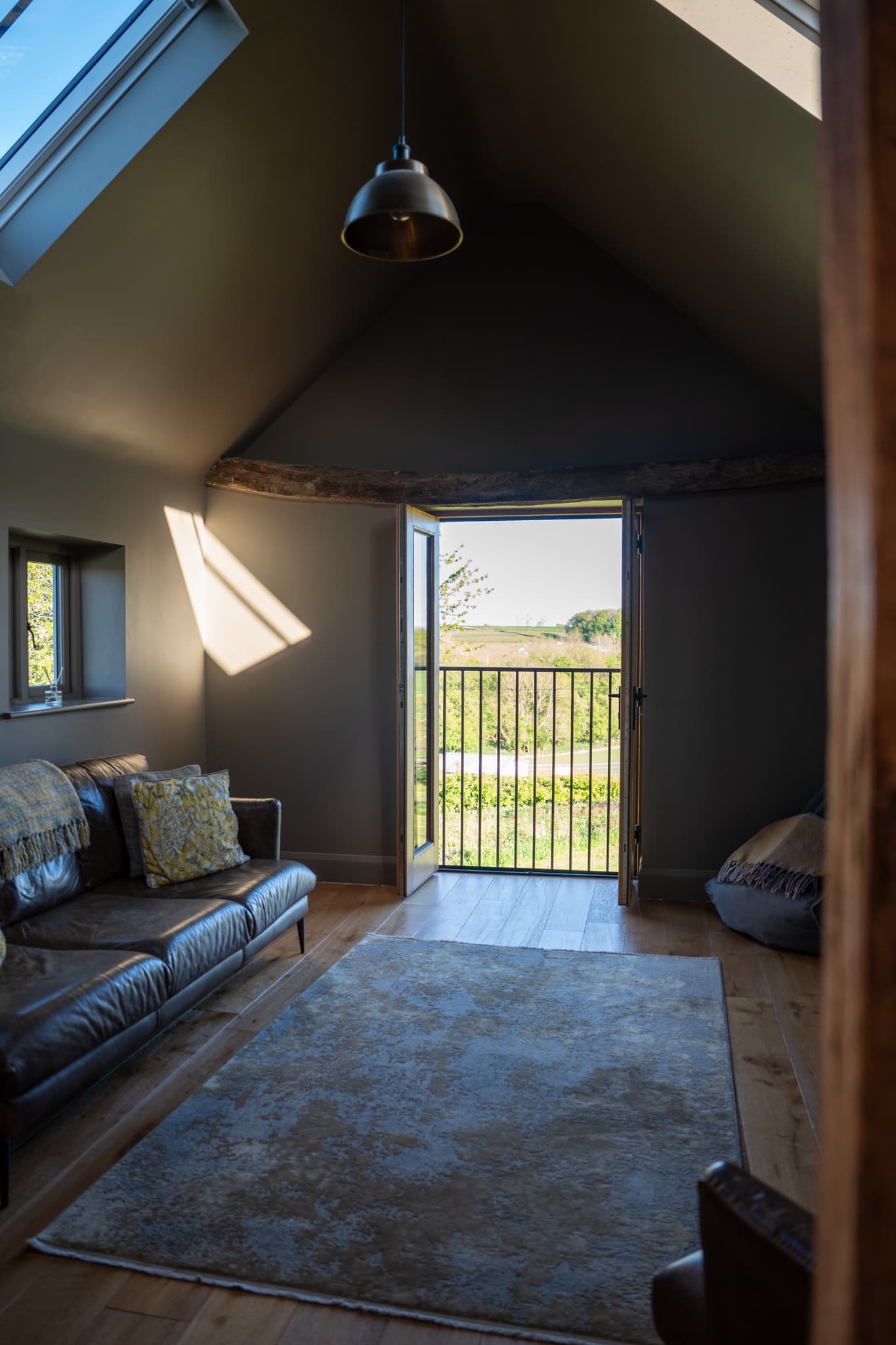 The first-floor sitting room with the Juliet balcony doors standing open onto a hedge-lined meadow. A pendant light hangs from the vaulted ceiling, a rooflight throws sun across the wall, and a leather sofa and rug sit under the original curved oak beam.