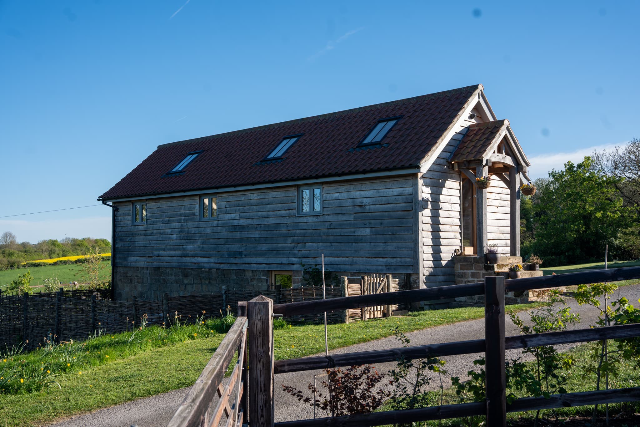 Orchard Barn seen along its length from the farm track on a clear spring morning: silvery weathered timber cladding, a dark tiled roof set with three rooflights, the oak porch at the far end, and post-and-rail fencing across the foreground.