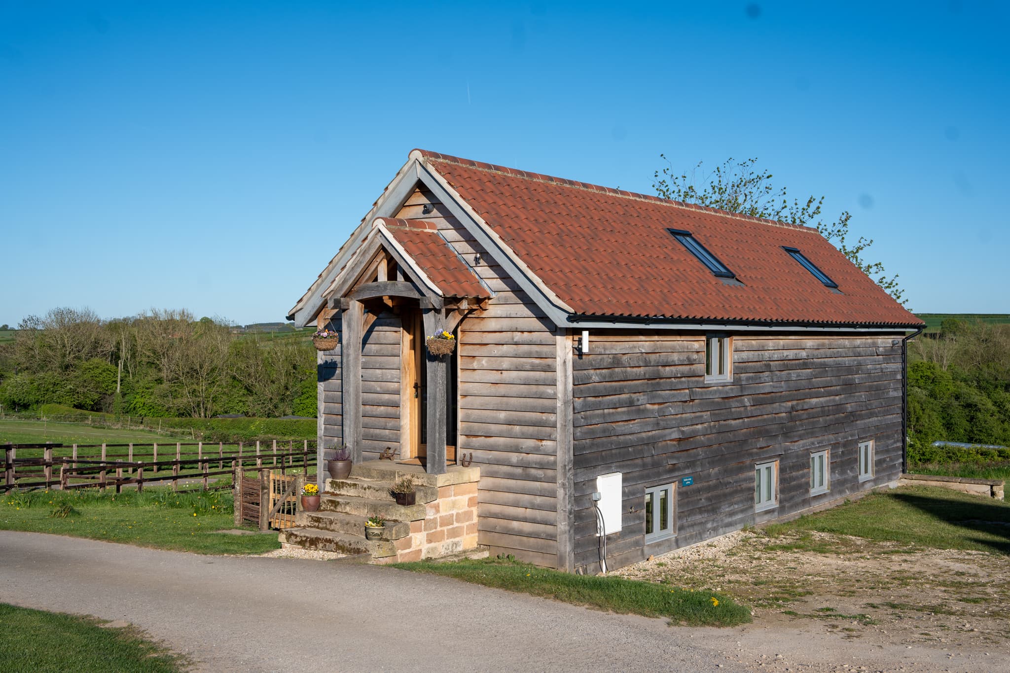 Orchard Barn from the gravel lane — an oak porch over the front door, stone steps up to it, red pantile roof and timber-clad walls, with open fields and hedgerow behind under a cloudless blue sky.