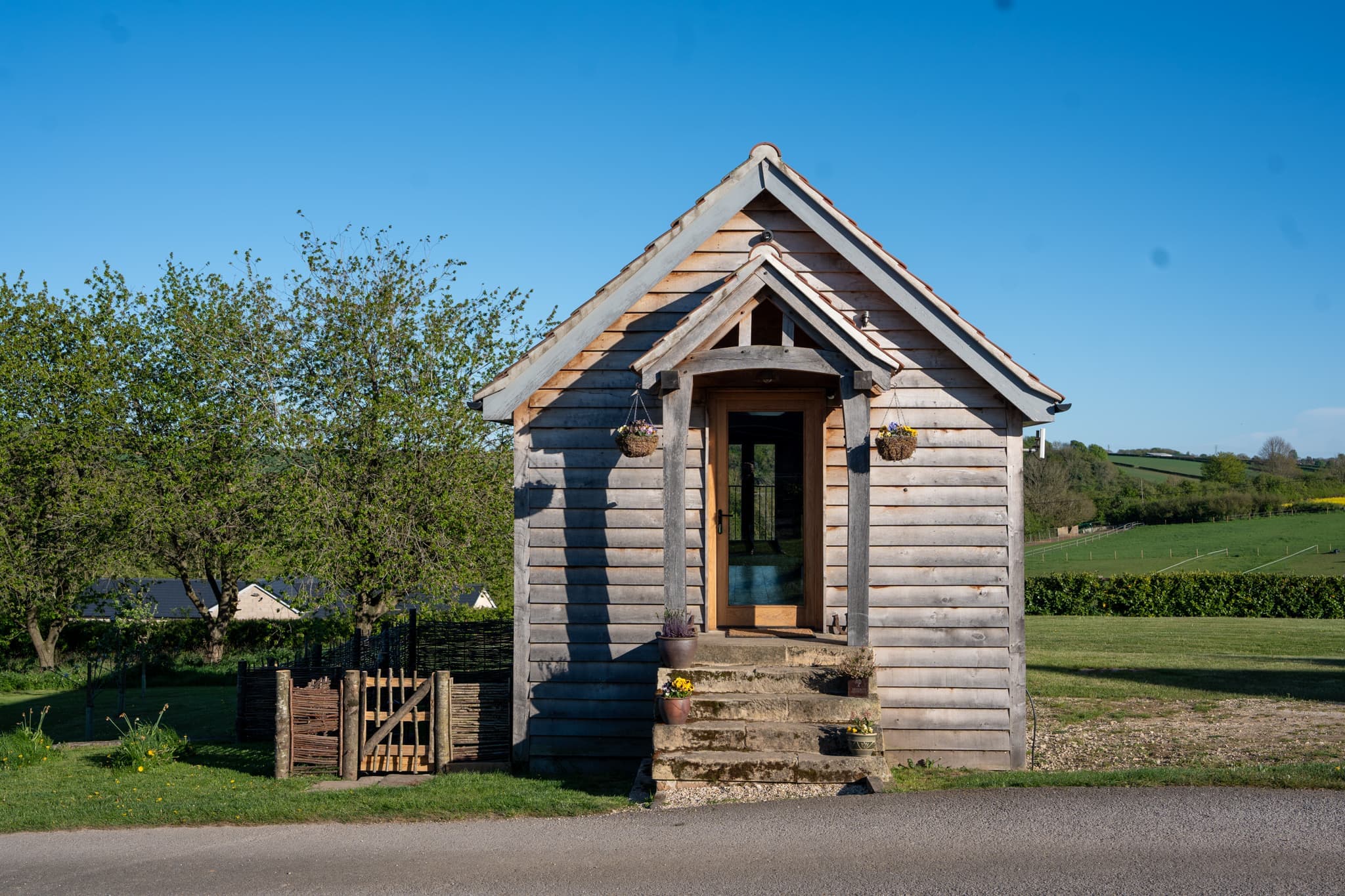 The gable end of Orchard Barn: an oak porch with a hanging basket either side of the glazed front door, stone steps down to a wooden gate, and the farm lane running past mown grass and orchard trees.