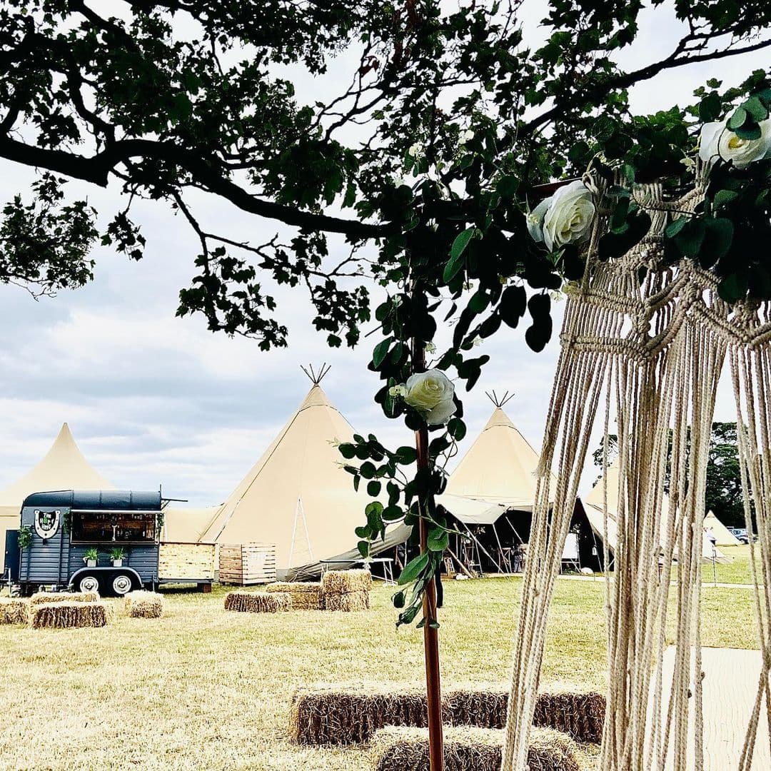 A restored vintage horsebox bar parked on grass beside straw bales, with tipis and a flower-dressed macrame arch in the foreground
