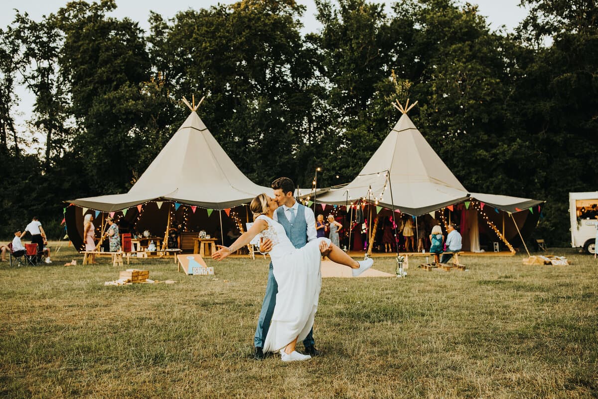 A couple dancing on the grass at dusk in front of two giant linked tipis strung with bunting and festoon lights, guests inside